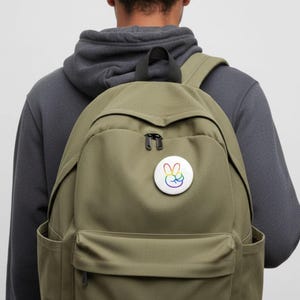 May include: An olive green backpack with a front pocket and a black zipper. A white button with a rainbow peace sign is pinned on the front. The person is wearing a gray hoodie.