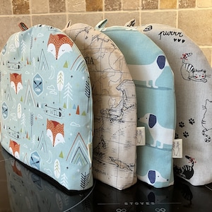 May include: Four teal blue, grey, and beige fabric tea cosy covers with different patterns. The covers feature a fox, a map, a dachshund, and a cat. The text "purrr" is on the cat cover. The text "Summer Camp" is on the fox cover.