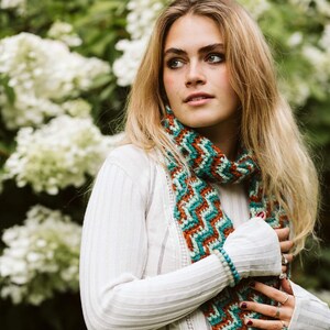 May include: A woman wearing a white top and blue jeans, with a patterned scarf. The scarf has a zig-zag design in teal, orange, and white. The background features white flowers. The scarf is a key feature of the outfit.