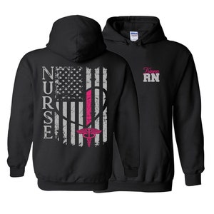 May include: Black hooded sweatshirt with a distressed American flag design and the word "NURSE" printed vertically on the back. The flag has a pink heart with a medical caduceus symbol. The front of the sweatshirt has the name "Triana RN" printed in white.