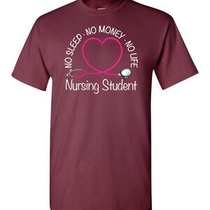 Peut inclure: T-shirt bordeaux avec un motif de st&eacute;thoscope rose en forme de c&oelig;ur. Le texte indique "NO SLEEP - NO MONEY - NO LIFE" et "Nursing Student" en blanc. Un v&ecirc;tement d&eacute;contract&eacute; pour les &eacute;tudiants en soins infirmiers.