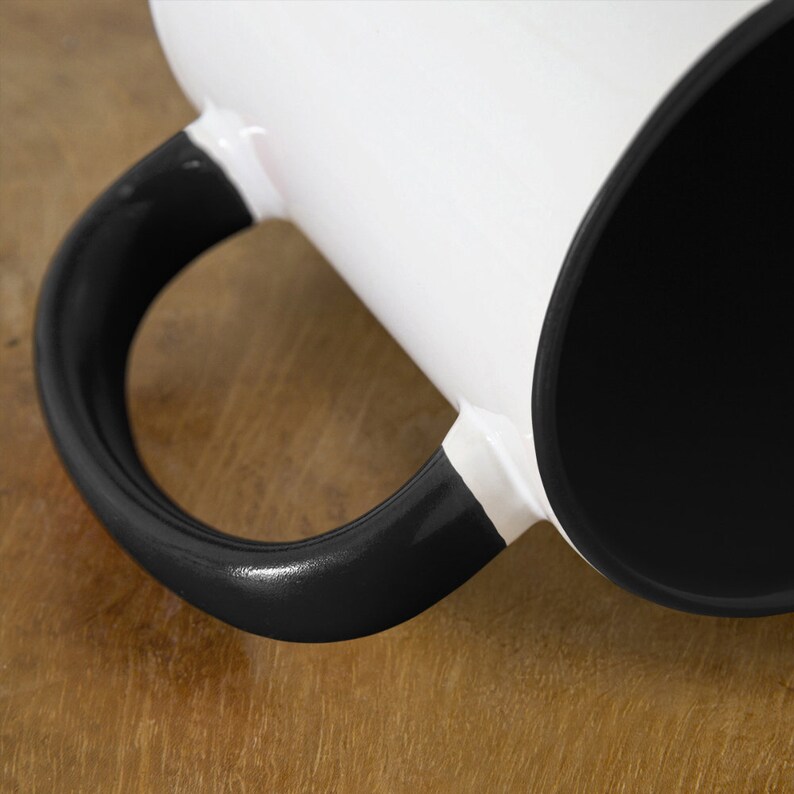 May include: A white ceramic mug with a black handle and a black interior. The mug is sitting on a wooden surface.