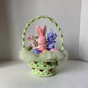 May include: A green crocheted Easter basket with a pink bunny, purple and white flowers, and pastel colored eggs.