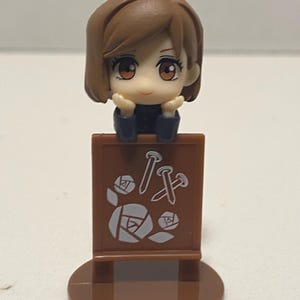 May include: A brown figurine of a character with brown hair and large eyes, resting their chin on their hands. The figurine is attached to a brown sign with white nail and rose graphics.