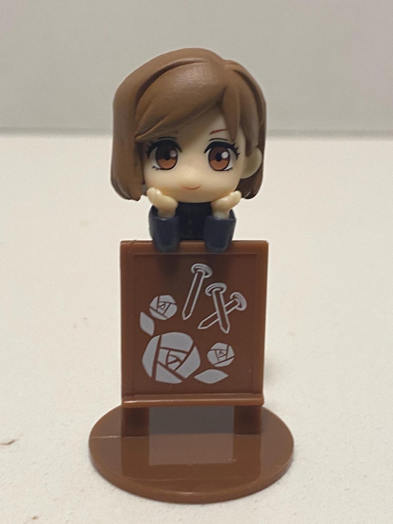 May include: A brown figurine of a character with brown hair and large eyes, resting their chin on their hands. The figurine is attached to a brown sign with white nail and rose graphics.
