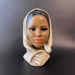 May include: A ceramic bust of a woman with dark skin, black hair, and orange lipstick. She wears a cream-colored head covering. The bust is a decorative item, likely for display.