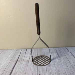 May include: A potato masher featuring a dark brown wooden handle attached to a silver-colored metal frame. The base consists of a circular metal plate with a grid of square cutouts designed for mashing food items in a kitchen setting.