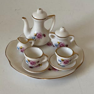 May include: A miniature porcelain tea set with a teapot, sugar bowl, creamer, and two cups and saucers. The set is white with gold trim and floral designs in pink, blue, and yellow. The set sits on a scalloped tray.