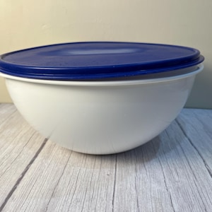 May include: A white mixing bowl with a smooth, rounded base and a wide rim. The bowl features a deep, circular design and is topped with a solid, dark blue plastic lid that includes a small, protruding tab on one side for easy removal.
