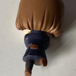 May include: A small figurine of a person with brown hair, wearing a dark blue coat and brown shoes. The figurine has a small brown satchel at the waist. The figure is posed as if running or walking.