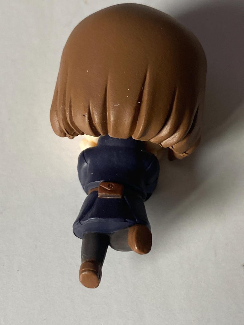 May include: A small figurine of a person with brown hair, wearing a dark blue coat and brown shoes. The figurine has a small brown satchel at the waist. The figure is posed as if running or walking.
