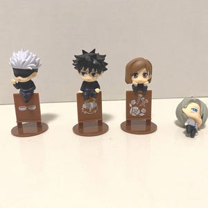 May include: Four small anime character figurines. Three figures are standing behind brown signs, and one is lying down. The figures have black outfits, and different hair colors, including white, black, and brown. The signs have illustrations.