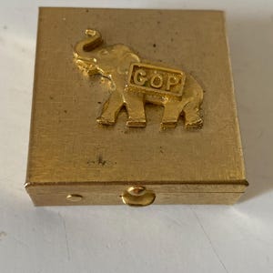 Puede incluir: Una caja rectangular dorada con un emblema de elefante en relieve y las letras "GOP" en un marco rectangular. La caja tiene una tapa con bisagras y un peque&ntilde;o cierre.