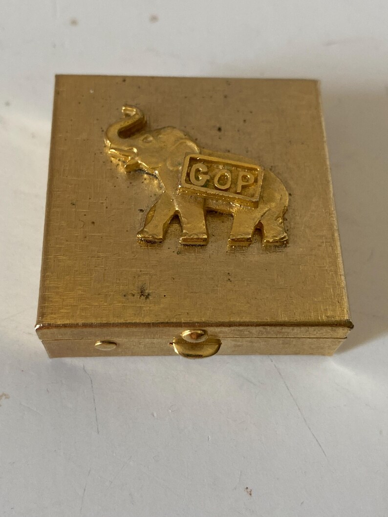 Puede incluir: Una caja rectangular dorada con un emblema de elefante en relieve y las letras "GOP" en un marco rectangular. La caja tiene una tapa con bisagras y un peque&ntilde;o cierre.