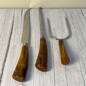 May include: A carving set with three pieces: a knife, a sharpening steel, and a carving fork. The blades and fork are silver-colored, and the handles have a marbled amber and brown pattern. The set is displayed on a light wood surface.
