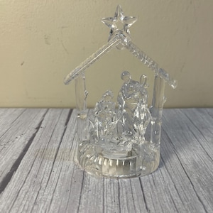 May include: A clear glass nativity scene figurine featuring a stable structure topped with a star. The piece depicts three figures standing and kneeling within the frame, all crafted from transparent, molded glass with a smooth, reflective finish.