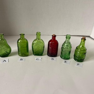 May include: Six small glass bottles with various designs and colors. The bottles are green, red, and clear. They are arranged in a row on a white surface.