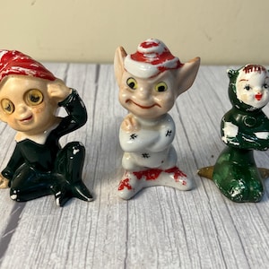 May include: Three ceramic figurines featuring a seated elf in a red hat and dark green suit, a standing elf with pointed ears wearing a white outfit with red accents, and a small figure in a dark green hooded suit with a painted face.