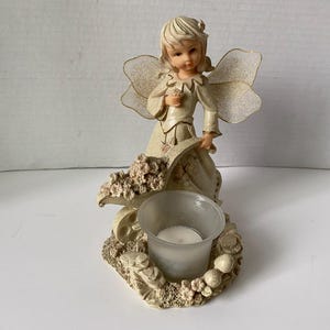May include: A white ceramic angel figurine with wings holding a flower, standing on a base with a tea light holder. The base is decorated with flowers and leaves.