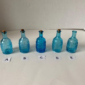 May include: Five small, blue glass bottles with cork stoppers. The bottles are all different shapes and sizes, but they all have a similar textured surface. The bottles are labeled A through E.