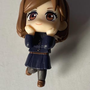 May include: A small figurine of a female anime character with brown hair and eyes. She wears a dark blue school uniform with a pleated skirt and brown shoes. The figure is posed with hands near her face, smiling.