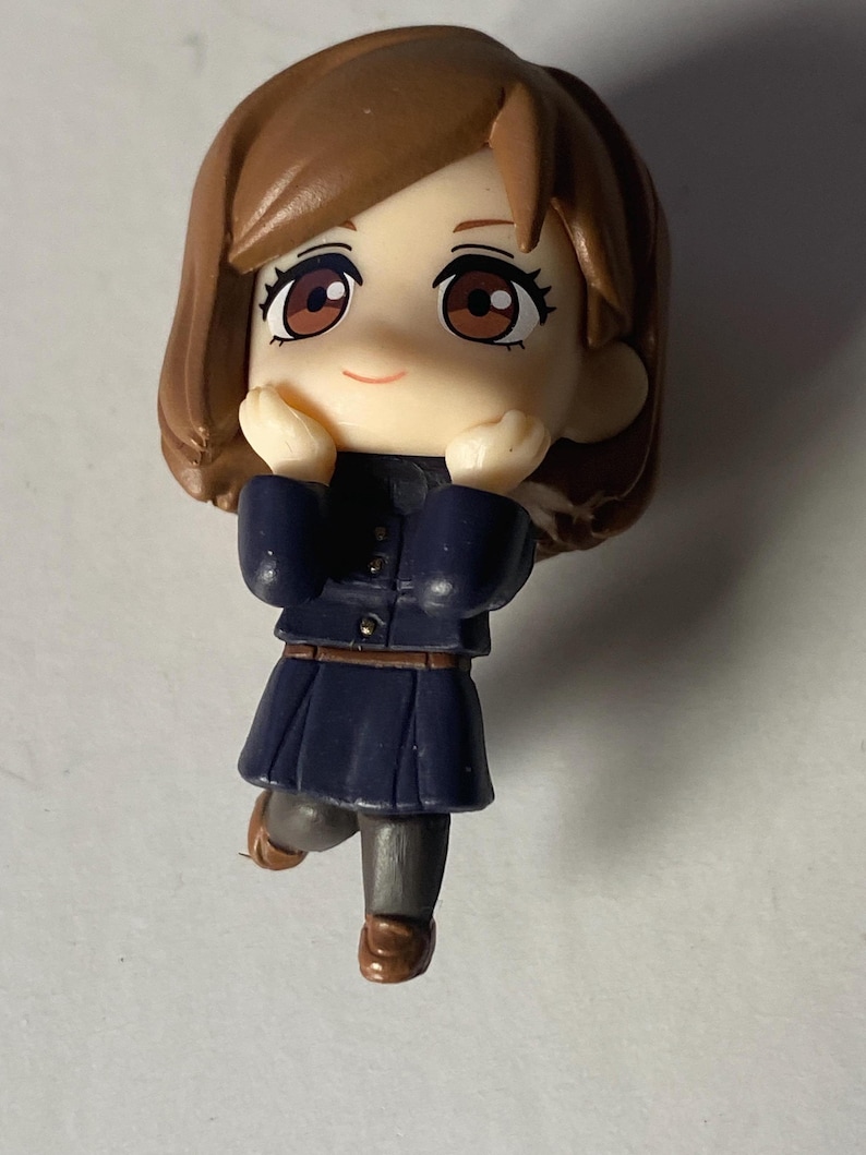 May include: A small figurine of a female anime character with brown hair and eyes. She wears a dark blue school uniform with a pleated skirt and brown shoes. The figure is posed with hands near her face, smiling.