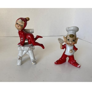 May include: Two vintage ceramic figurines. One is a red-suited elf on a white stool with gold accents. The other is a chef elf in a white hat and red suit, holding a plate. Both have detailed facial features.