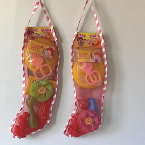 May include: Two Christmas stockings filled with vintage toys. Each stocking is made of red mesh with a pink and white striped trim and handle. The toys include scissors, a spinning top, and other colorful plastic items.