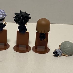 May include: Four small character figurines. Three figures stand on brown rectangular bases with circular bottoms. The figures have black outfits and different hairstyles. One figure is lying down on the surface.