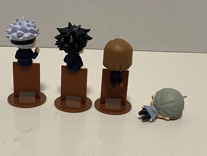 May include: Four small character figurines. Three figures stand on brown rectangular bases with circular bottoms. The figures have black outfits and different hairstyles. One figure is lying down on the surface.