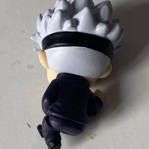 May include: A small figurine of a character with spiky white hair, a black blindfold, and a dark blue outfit. The figure is in a running pose, with the back of the head and body visible.