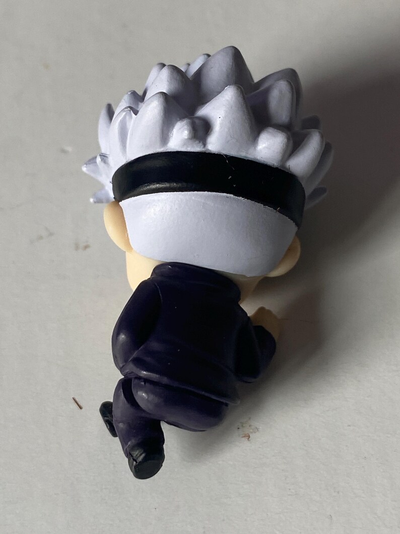 May include: A small figurine of a character with spiky white hair, a black blindfold, and a dark blue outfit. The figure is in a running pose, with the back of the head and body visible.