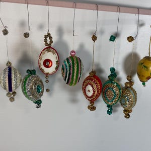 May include: Eight decorative hanging ornaments suspended from a pink bar. The collection includes various egg-shaped designs featuring intricate beadwork, sequins, metallic accents, and colorful patterns in shades of red, green, blue, and gold.