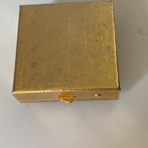 Puede incluir: Una peque&ntilde;a caja rectangular dorada con una superficie texturizada. La caja tiene una tapa con bisagras y un peque&ntilde;o cierre en la parte delantera. La caja parece ser de metal y tiene un aspecto ligeramente desgastado.