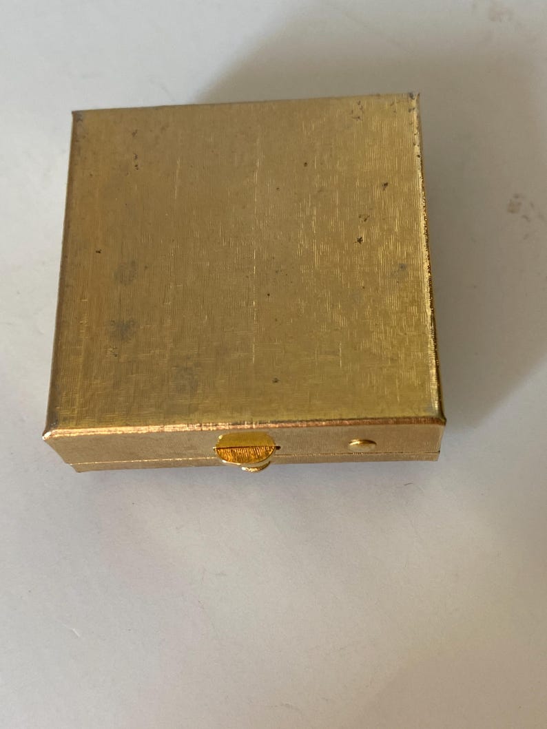Puede incluir: Una peque&ntilde;a caja rectangular dorada con una superficie texturizada. La caja tiene una tapa con bisagras y un peque&ntilde;o cierre en la parte delantera. La caja parece ser de metal y tiene un aspecto ligeramente desgastado.