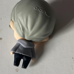 May include: A small figurine of a person with gray hair, wearing a gray jacket and dark blue pants. The figurine has a silver loop at the top. The text "&copy;HTB/YDP" is printed on the back of the head.
