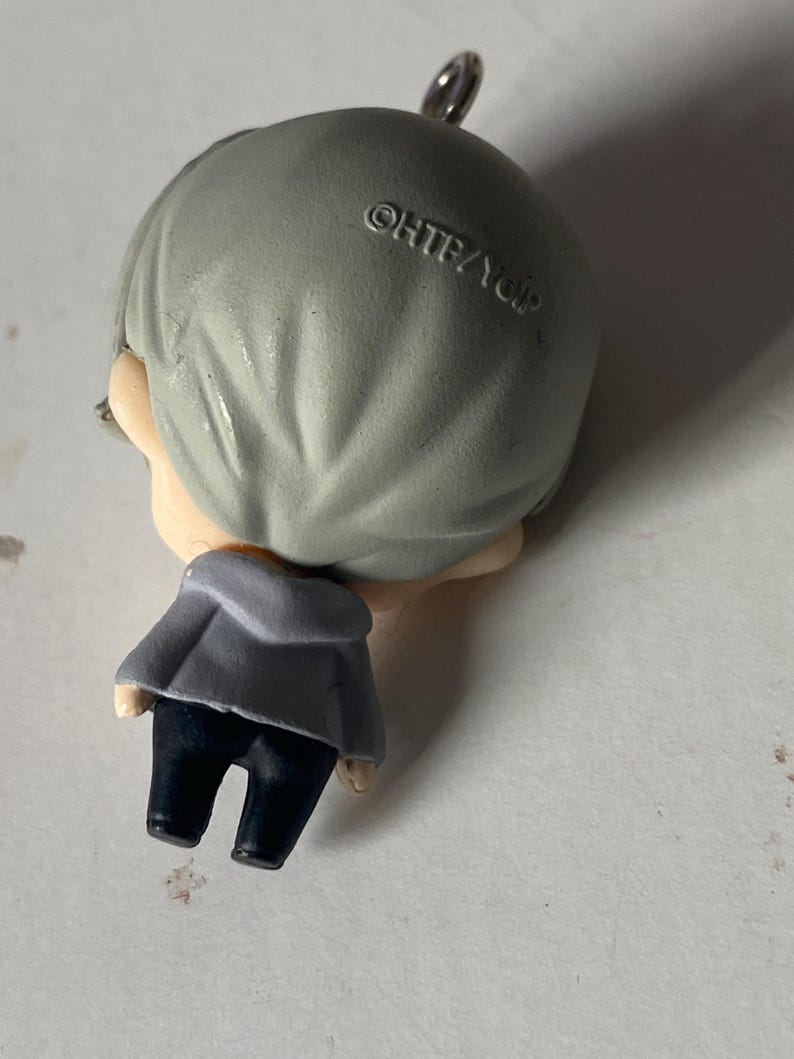 May include: A small figurine of a person with gray hair, wearing a gray jacket and dark blue pants. The figurine has a silver loop at the top. The text "&copy;HTB/YDP" is printed on the back of the head.