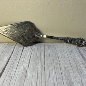 May include: Silver-toned cake server with an ornate, floral-patterned blade and a detailed handle. The server is designed for serving cakes and pies. The handle has a decorative, textured design. The server is resting on a wooden surface.