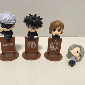 May include: Four small anime character figurines. Three figures are seated on brown display stands. The first figure has white hair and a black blindfold. The second figure has black spiky hair. The third figure has brown hair. The fourth figure is lying down.