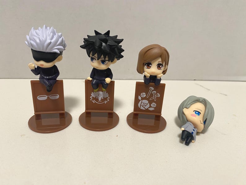 May include: Four small anime character figurines. Three figures are seated on brown display stands. The first figure has white hair and a black blindfold. The second figure has black spiky hair. The third figure has brown hair. The fourth figure is lying down.