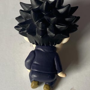 May include: A small figurine of a character with spiky black hair, wearing a dark blue jacket and brown shoes. The figurine is viewed from the back, showcasing the details of the hair and clothing.
