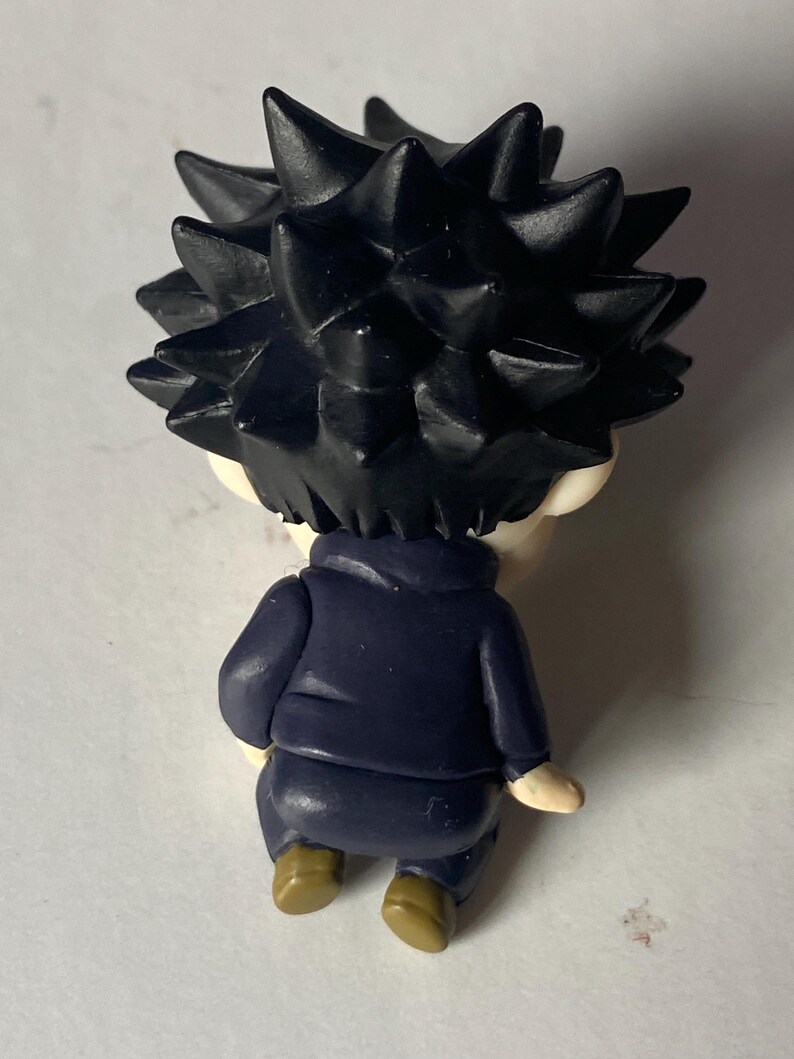 May include: A small figurine of a character with spiky black hair, wearing a dark blue jacket and brown shoes. The figurine is viewed from the back, showcasing the details of the hair and clothing.