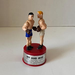 May include: A vintage toy featuring two small boxer figures facing each other, mounted on a red base. The boxers wear gloves and shorts. The base has text that reads "HIT AND MISS PUSH BUTTON PUPPET".