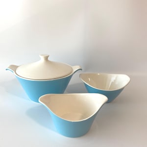 May include: A set of three ceramic serving dishes. The set includes a lidded bowl and two smaller bowls. The exterior of the bowls is light blue, and the interior is off-white. The lidded bowl has handles on either side.