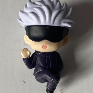 May include: A small figurine of a character with white spiky hair, a black blindfold, and a dark purple outfit. The figurine is in a seated position with one arm raised. The figure has a neutral expression.