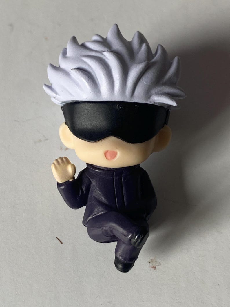 May include: A small figurine of a character with white spiky hair, a black blindfold, and a dark purple outfit. The figurine is in a seated position with one arm raised. The figure has a neutral expression.