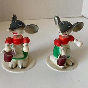 May include: Two vintage mouse figurines. Each mouse has a gray head, large ears, and is wearing a red and green outfit. They are holding a small red bucket. The figurines are standing on a white base.