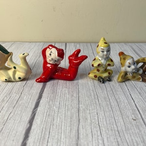 May include: Four small ceramic elf figurines in various poses. One is cream with green spots and a green hat, one is solid red, one is yellow with brown spots in a seated position, and one is brown and yellow with a pointed hat, all with painted faces.