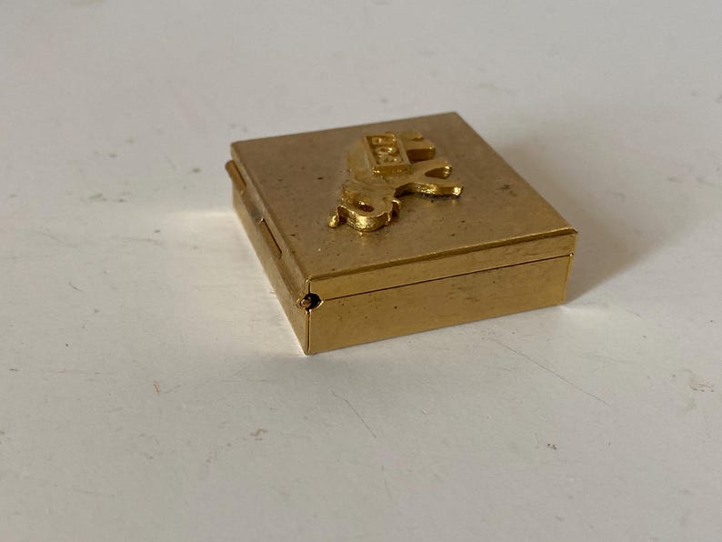 Puede incluir: Peque&ntilde;a caja rectangular dorada con una figura de caballo en relieve en la tapa. La caja tiene un dise&ntilde;o con bisagras y parece estar hecha de metal. La figura del caballo tambi&eacute;n es dorada y a&ntilde;ade un elemento decorativo.
