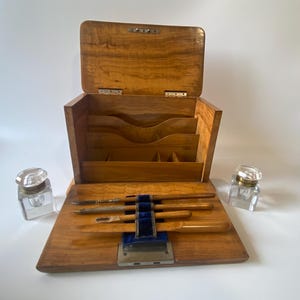 May include: A wooden desk organizer with a hinged lid and interior compartments for stationery. The front panel features a blue velvet holder containing four writing instruments. Two clear glass inkwells with silver-toned caps sit on either side.